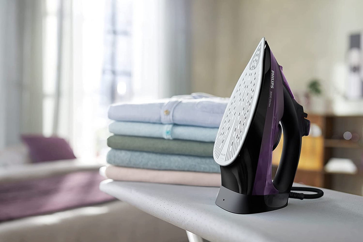 Philips DST5030/80 5000 Series steam iron standing on its heel rest on an ironing board beside a stack of folded shirts