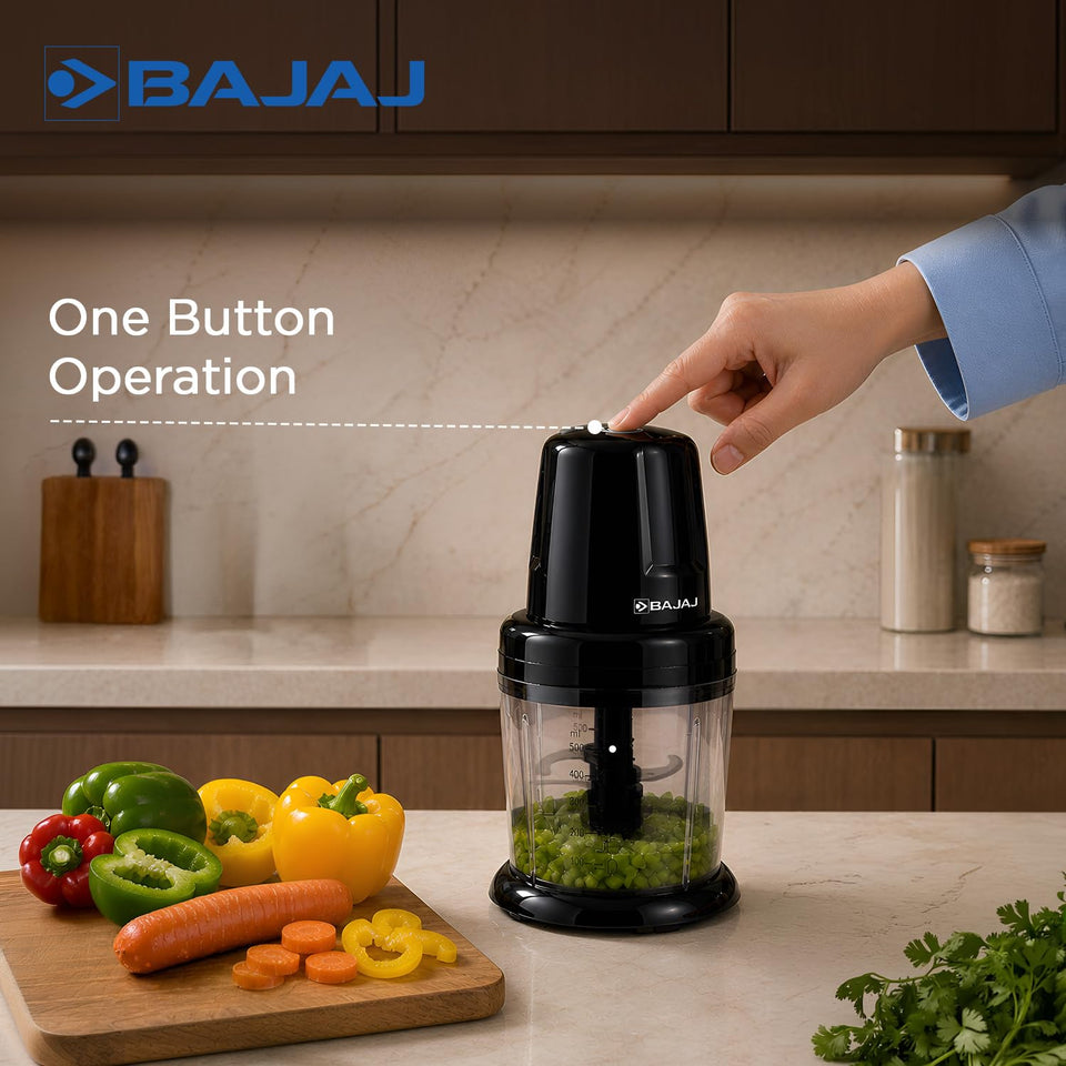 Hand pressing the single top button of the Bajaj Chop Pro chopper, with chopped green vegetables in the bowl and bell peppers and carrot nearby