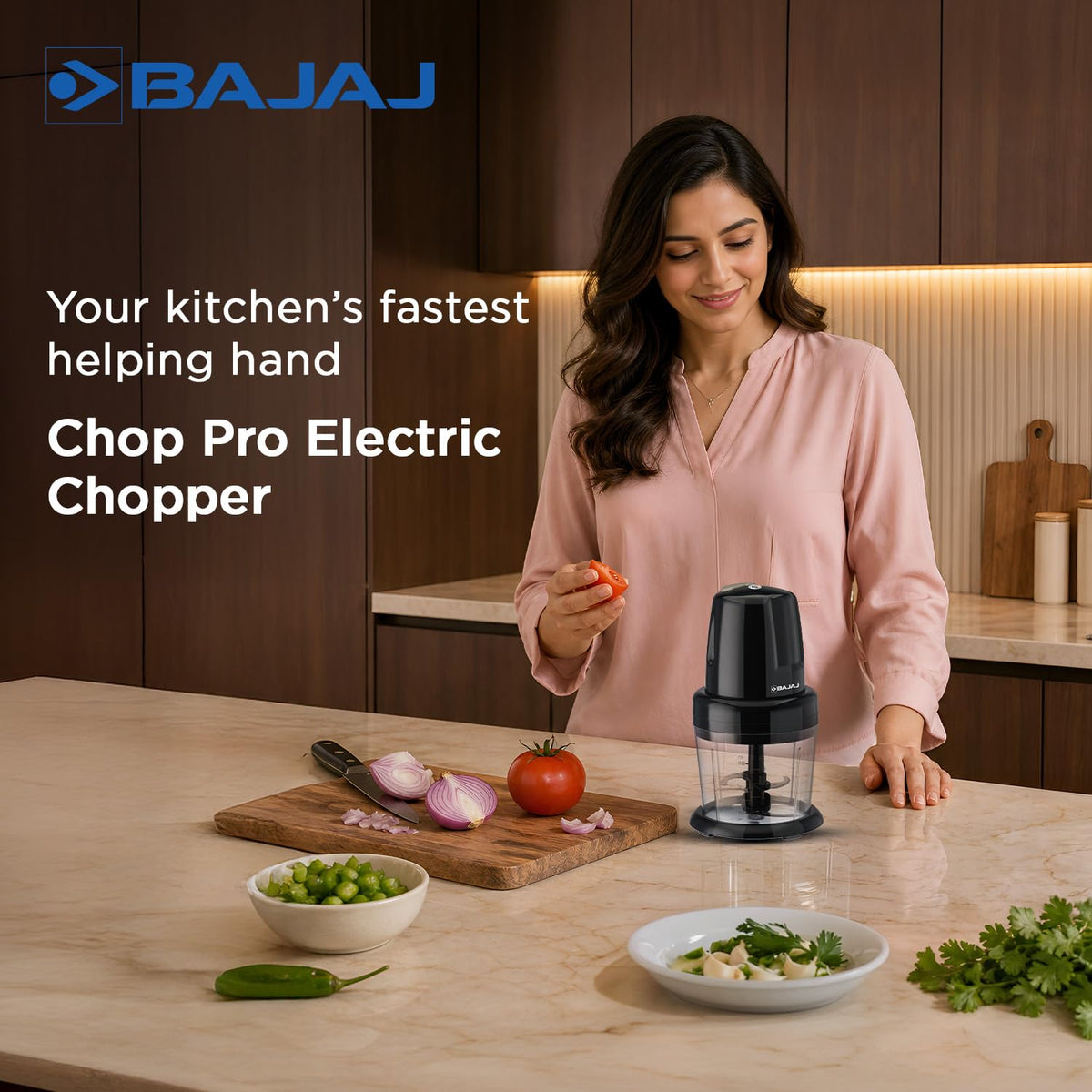 Bajaj Chop Pro electric chopper on a kitchen counter beside a chopping board with onion, tomato and green chillies, with a woman holding a tomato