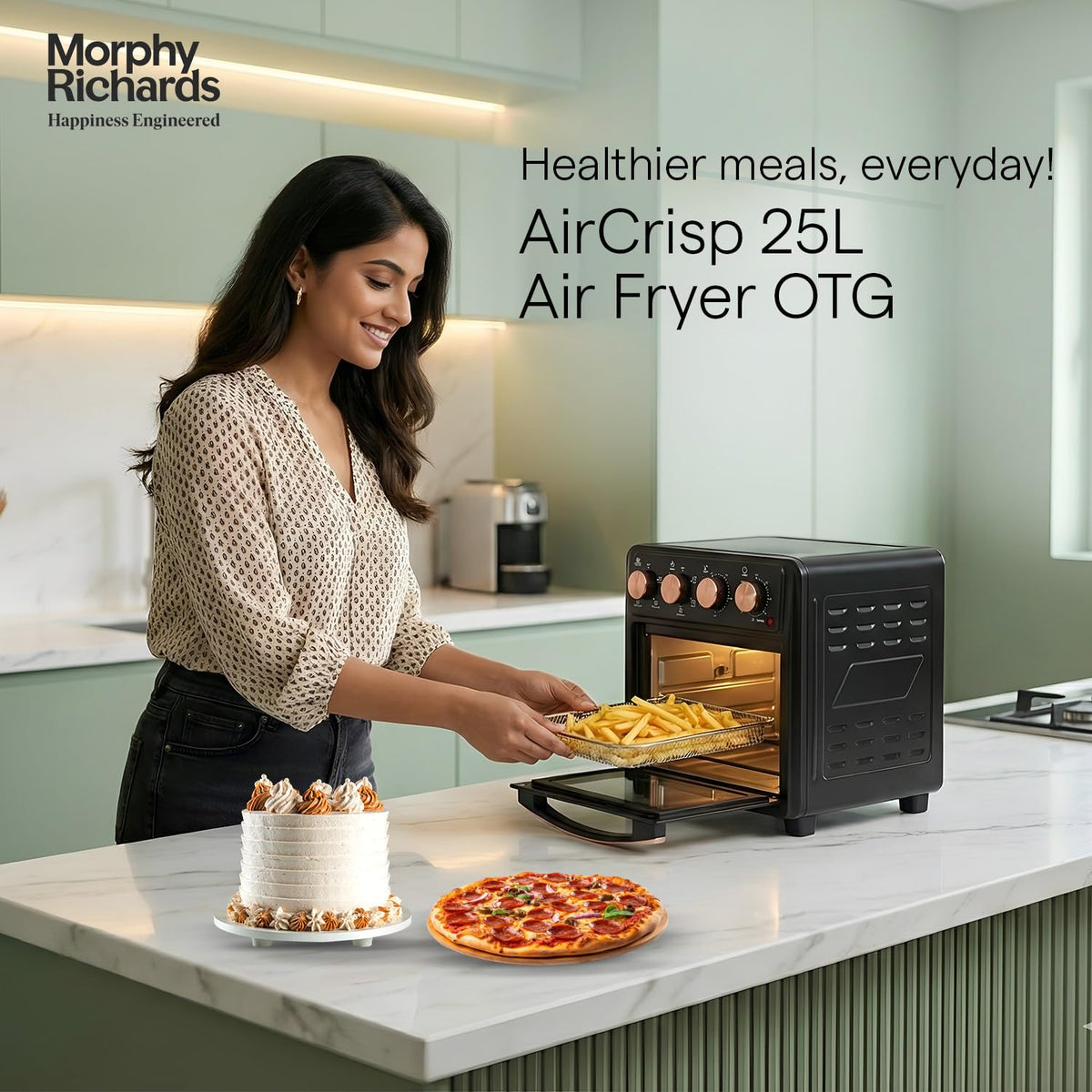 Woman placing a basket of fries into the Morphy Richards 510060 AirCrisp 25 L air fryer oven on a kitchen counter, with a cake and pizza in front